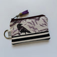 A fabric zip pouch with a raven and branch pattern in black and muted purple on a cream background, featuring black and white horizontal stripes on the bottom, a purple tassel, and a metal key ring with a tag reading 'BEEGO HANDCRAFTED'.