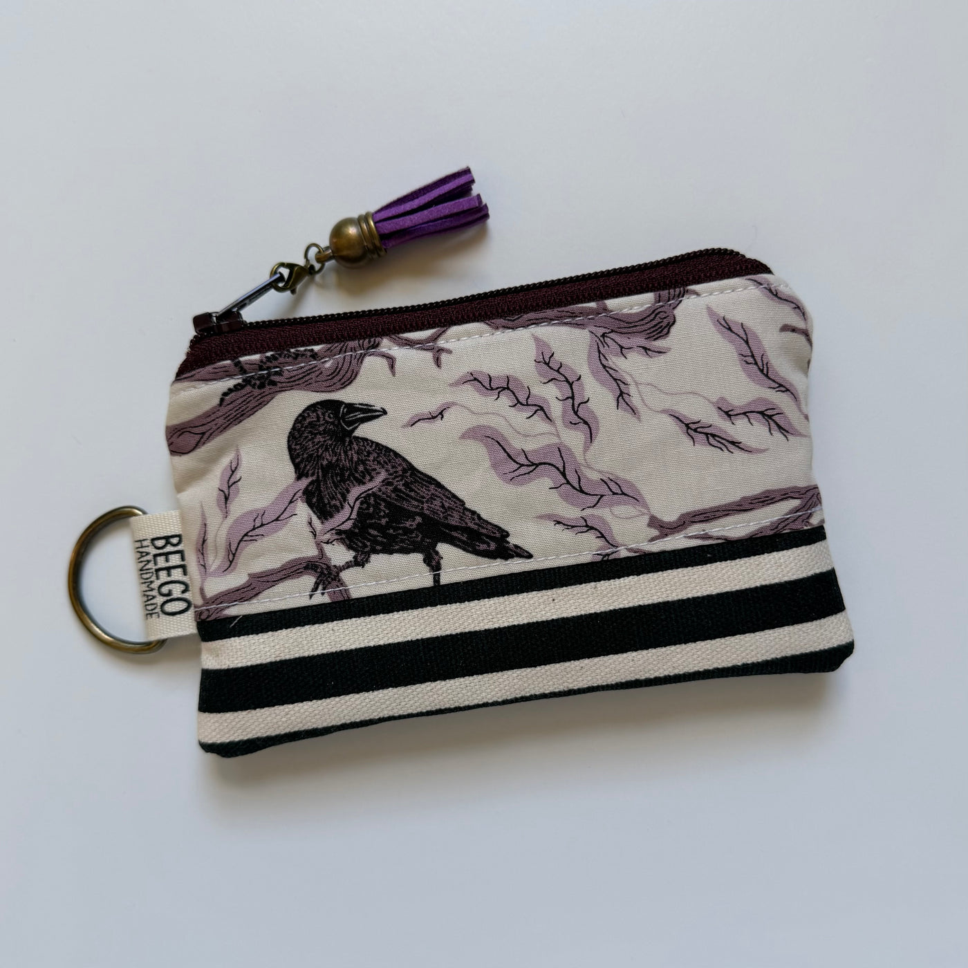 A fabric zip pouch with a raven and branch pattern in black and muted purple on a cream background, featuring black and white horizontal stripes on the bottom, a purple tassel, and a metal key ring with a tag reading 'BEEGO HANDCRAFTED'.